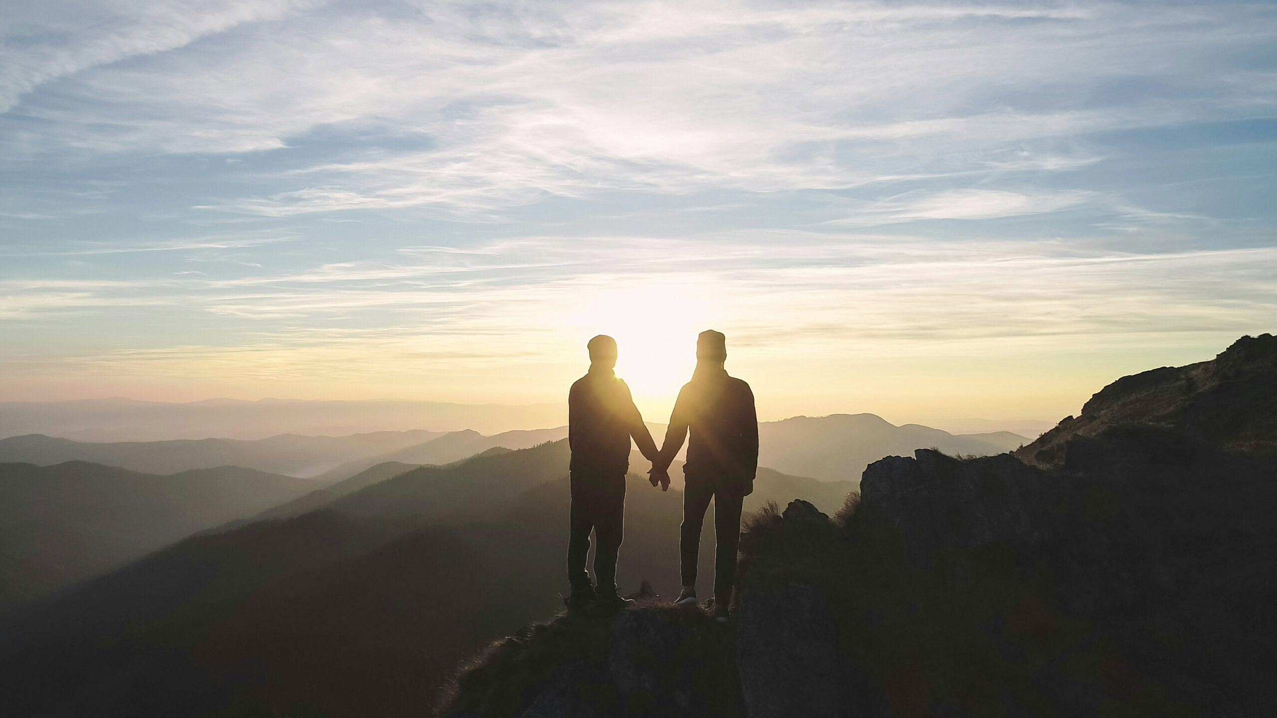 2 personnes se tiennent la main au sommet d'une montagne face au soleil levant